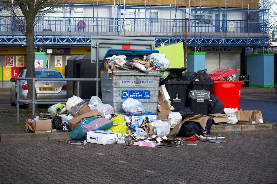 A cluttered waste disposal area on a paved sidewalk in front of a commercial building with a blue metal facade and large windows, featuring multiple overflowing black, white, and red garbage bins fill